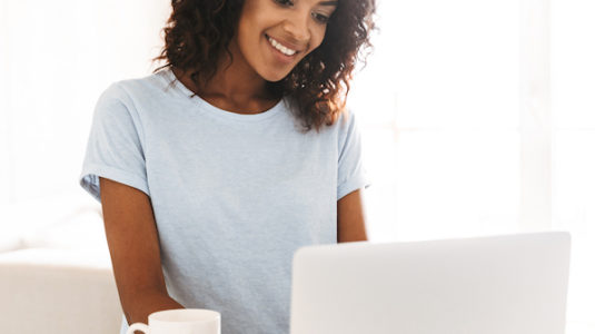 Smiling african woman using laptop computer while sitting at home with cup of coffee